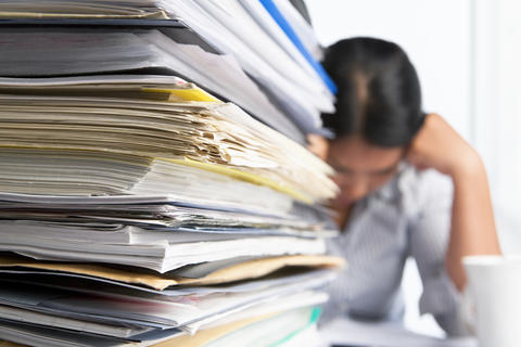 Large stack of papers and files on a desk, with a woman sitting behind them with her head in her hands.