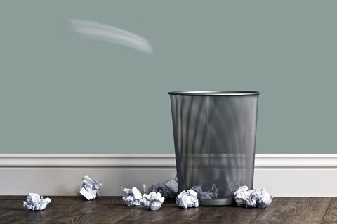 Mesh trash can against a wall with wads of paper surrounding it on the floor.